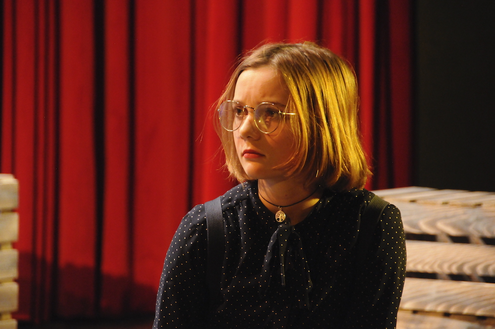 Blondes Mädchen mit Brille, im Hintergrund Theatervorhang