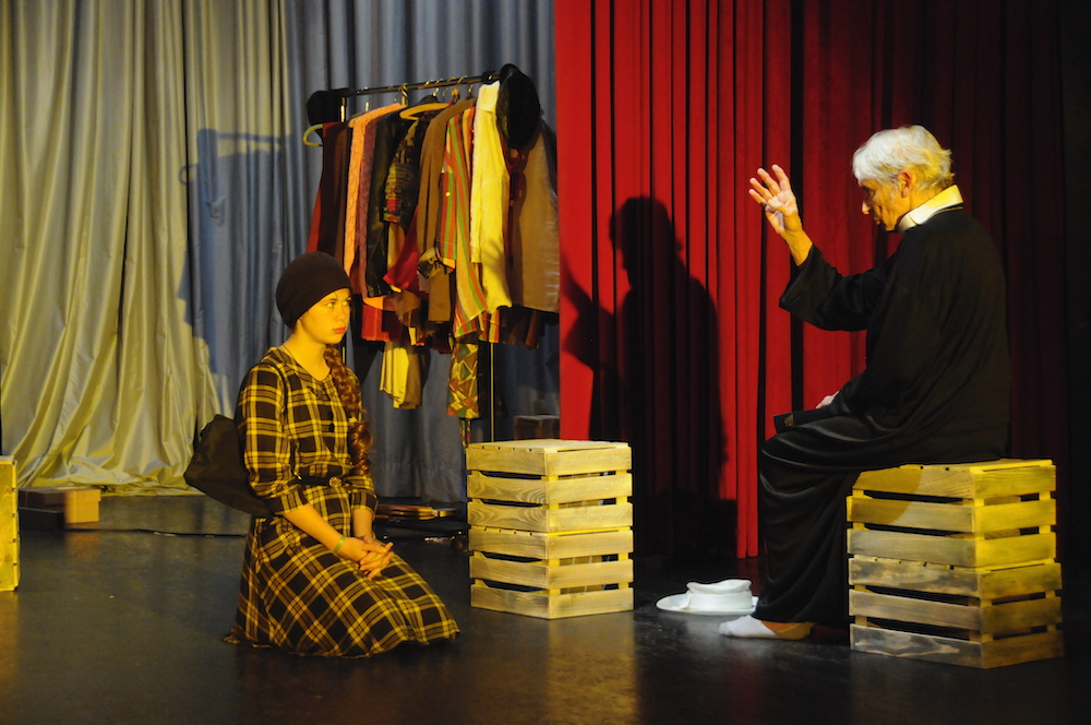 Auf einer Bühne: Mädchen im Karokleind sitzt vor segnendem Pfarrer